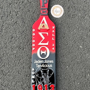 May include: A red, black, and gray wooden paddle with white and gold lettering. The paddle features the Greek letters Delta Sigma Theta, the name "Jaden Jones Ten4cious", and an illustration of two elephants. The year 1913 is also displayed.