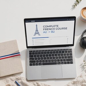 Peut inclure: Un ordinateur portable argenté affichant un cours complet de français A2 à B2 avec une icône de la tour Eiffel, accompagné d'un carnet beige avec un ruban bleu, blanc et rouge, un casque noir, une tasse de café et un petit drapeau tricolore.