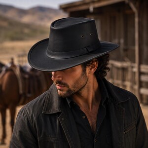 May include: A black leather fedora-style hat with a wide brim, a structured crown, and a matching leather hat band. The hat features two small metal ventilation eyelets on each side of the crown, providing a clean and functional design.