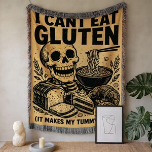 May include: A woven throw blanket in yellow with black graphics featuring a skull, a bowl of noodles, bread, and cake. The text reads I CANT EAT GLUTEN and IT MAKES MY TUMMY. The blanket has a fringed edge with multicolored threads.