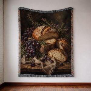 May include: A woven throw blanket featuring a still life print of crusty sourdough bread loaves, dark purple grapes, and sprigs of green rosemary on a wooden surface, finished with decorative fringed edges on the top and bottom sides.