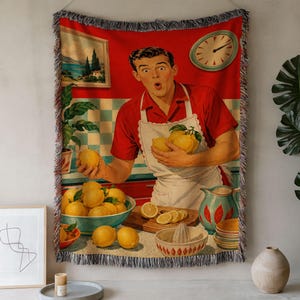 May include: A woven throw blanket featuring a vintage-style illustration of a person in a red shirt and white apron holding lemons in a kitchen. The scene includes a bowl of lemons, a juicer, a pitcher, and a wall clock on a red and tiled background.