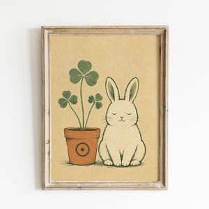 May include: A framed wall art print on a tan background featuring a white rabbit with closed eyes sitting next to a terracotta-colored pot containing a green three-leaf clover plant. The pot has a circular design on its front surface.
