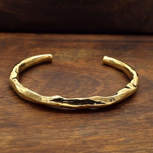 Könnte beinhalten: Ein goldfarbenes Armreif-Armband mit strukturierter, gehammerter Optik. Das offene Design weist eine unregelmäßige, organische Form auf, die auf ein handgefertigtes Stück hindeutet. Das Armband wird auf einer Holzoberfläche präsentiert.