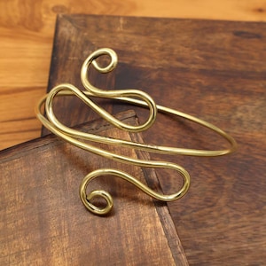 Op de afbeelding: Een goudkleurige armmanchet met een open ontwerp. De manchet heeft een wervelend, abstract patroon met afgeronde uiteinden. De sieraden worden gepresenteerd op een donker houten oppervlak.
