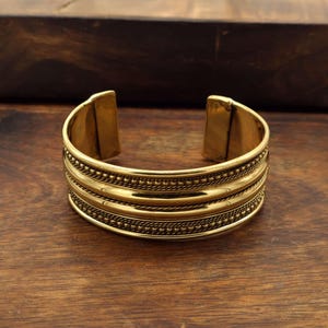 Puede incluir: Un brazalete de puño dorado con un diseño de banda ancha. El brazalete presenta múltiples bandas texturizadas con una combinación de patrones de cuentas y cuerdas. El diseño abierto permite un uso fácil. El brazalete se muestra sobre una superficie de madera.