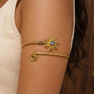 May include: A gold-toned metal arm cuff featuring an open-ended design with a spiral terminal on one side and a sun-shaped motif on the other. The sun emblem is centred with a small, round, iridescent blue stone set within its textured rays.