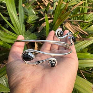 Puede incluir: Un brazalete plateado con detalles de piedras preciosas negras. El brazalete presenta un diseño en espiral con tres piedras negras y detalles decorativos. La joya se muestra sobre un fondo de follaje verde.
