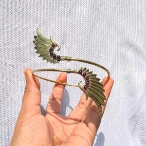 Può includere: Un bracciale da braccio in metallo color oro con due estremità simmetriche a forma di ala. Ogni ala è decorata con una fila di piccole pietre preziose rotonde rosso scuro incastonate lungo la curva interna della struttura metallica.