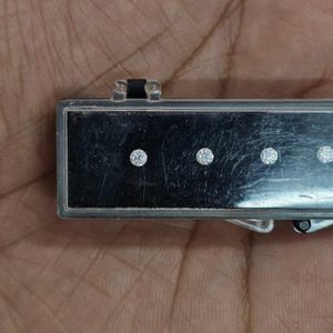 May include: A rectangular, silver-toned jewellery case with a black interior. The case holds four round, sparkling gemstones. The case is held in a person's hand.