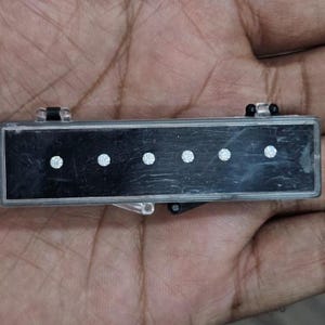 May include: A black rectangular object with five evenly spaced, circular, white embellishments. The object has a silver frame and clear plastic hinges. The item is held in a hand.