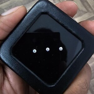 May include: A black leather case with a diamond-shaped window, displaying three small, sparkling diamonds. The diamonds are evenly spaced against a black backdrop, held in a person's hand.