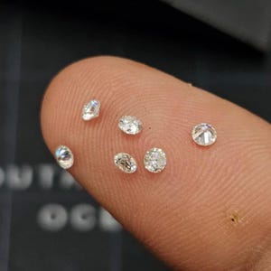 May include: A collection of small, sparkling diamonds of various shapes, including round and oval cuts. The diamonds are clear and reflect light, showcasing their brilliance. The gems are displayed on a finger, highlighting their size and clarity.