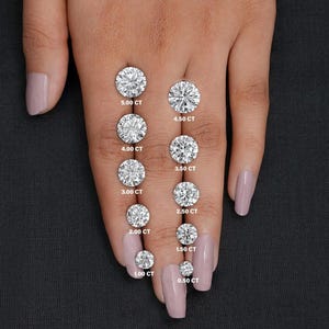May include: A hand shows a selection of round-cut diamonds, from 0.50 CT to 5.00 CT. Each diamond is labelled with its carat weight in white text. The diamonds are presented against a plain backdrop.