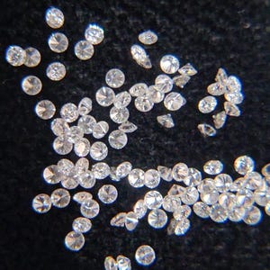 May include: A scattering of small, clear diamonds on a dark background. The diamonds are round, faceted, and reflect light, with slight variations in size and shape. The close-up image emphasizes the clarity and sparkle of the gemstones.