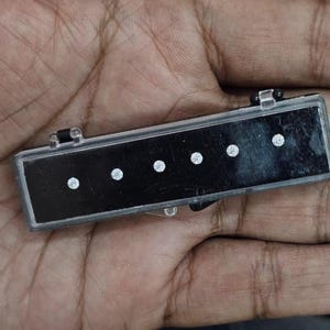 May include: A rectangular black jewellery case with a clear lid, containing five small, round, sparkling diamonds. The case is held in a hand, showcasing the gems.