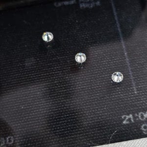 May include: Three small, round, clear gemstones are displayed on a dark, textured surface. The gemstones are faceted, reflecting light. The image is a close-up, highlighting the clarity and brilliance of the stones.