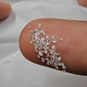 May include: A collection of small, sparkling diamonds. The diamonds are clear and faceted, reflecting light. They are scattered across a fingertip, showcasing their size and brilliance. The background is a soft, neutral color.