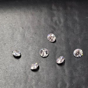 May include: Six clear, round-cut gemstones are displayed on a dark surface. The gemstones have multiple facets that reflect light, creating a sparkling effect. The gems are likely diamonds or diamond simulants.