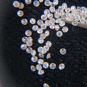 May include: A collection of small, round, clear diamonds scattered on a black surface. The diamonds are faceted, reflecting light and creating a sparkling effect. Some are held by tweezers.