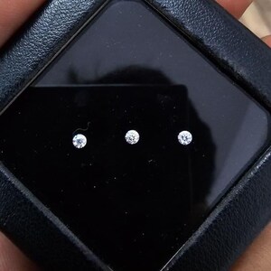 May include: Three small, round, sparkling diamonds are presented in a black, square case. The diamonds are set against a black background, and the case is held in a person's hand. The diamonds appear to be of high quality.