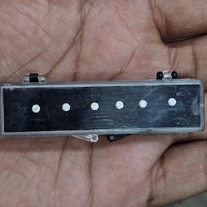 May include: A rectangular black component with five silver dots, held in a hand. The object has a clear plastic frame. The dots are evenly spaced along the length of the black surface. It appears to be a part of a larger device.