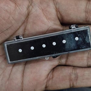 May include: A rectangular black case with a clear lid, containing five small, round, sparkling white gemstones. The case is held in a hand, with the gemstones evenly spaced along the black surface.