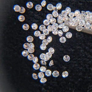 May include: A scattering of small, round, brilliant-cut diamonds on a dark background. The clear diamonds reflect light, highlighting their facets. Some are held by tweezers.