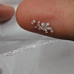 May include: A close-up shot shows a fingertip with a collection of small, clear diamonds. The diamonds are round and glitter, reflecting light. Some loose diamonds are scattered on a white surface below. The image focuses on the sparkle and clarity of the gemstones.