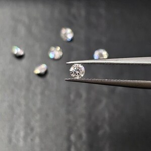 May include: Several small, round diamonds, one held by tweezers. The diamond held by the tweezers is in focus, displaying its facets. Other diamonds are scattered on a dark surface. The diamonds are clear and sparkling.