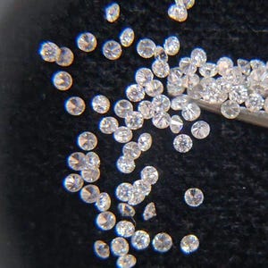 May include: A scattering of small, round, clear diamonds on a dark background. The diamonds are faceted, reflecting light, and vary slightly in size. Some are held by a metal tool.