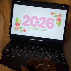 May include: A black tablet with a keyboard case displays a 2026 calendar with floral accents. The calendar includes sections for workout splits, hair and nail appointments, content, and expense tracking. The tablet is on a soft, textured surface.