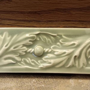 May include: A rectangular, pale green ceramic tile with a raised, ornate leaf and vine design. The tile features a central rope-like element and rounded accents on each end. The background is a dark wood.