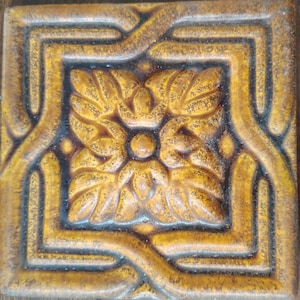 May include: Square, decorative tile with a raised floral design in the center. The tile is primarily a warm, golden-brown color with darker brown accents. The design features a central flower surrounded by stylized leaves and a border.
