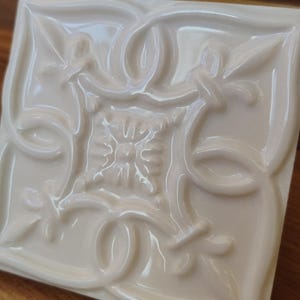 May include: A square, off-white ceramic tile with a raised, ornate floral design. The tile features a glossy finish and is set against a wooden surface. The design includes swirling patterns and stylized leaves.
