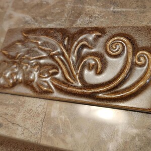 May include: Rectangular ceramic tile with a raised floral and scroll design. The tile features a blend of brown, tan, and cream colors. The raised design is highlighted with a darker brown glaze, creating a textured, decorative element.