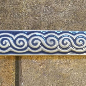 May include: A rectangular ceramic tile with a repeating wave pattern in shades of blue and white. The tile has a glossy finish and is likely used for decorative purposes in a kitchen or bathroom.