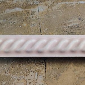 May include: A light pink ceramic tile with a rope-like design. The tile is rectangular and has a repeating pattern of curved lines. The tile is approximately 12 inches long.