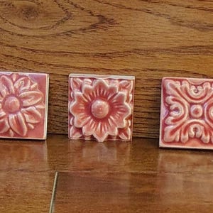 Puede incluir: Tres baldosas cuadradas de cerámica con diseños florales y ornamentales en relieve de color coral apagado. Cada baldosa presenta un patrón en relieve diferente, incluyendo un girasol y un rollo estilizado.