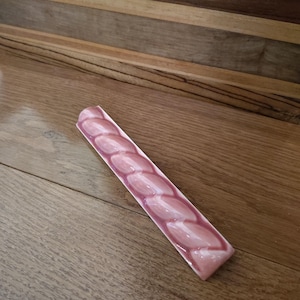 May include: A decorative, rectangular ceramic dish with a series of connected, oval-shaped indentations. The dish is primarily a light pink color with a darker pink border. The dish is resting on a wooden surface.
