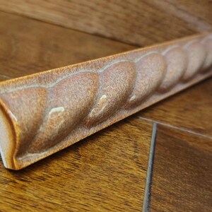 May include: A decorative architectural trim piece with a repeating rope-like design. The trim is a warm, earthy brown color with a glossy finish. The piece is rectangular and appears to be made of ceramic or a similar material.