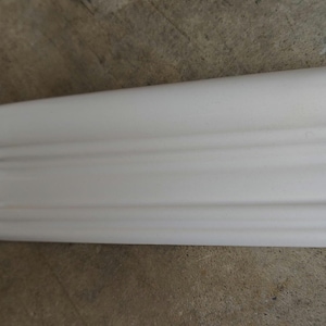 May include: A section of white ceramic molding with a curved top edge and a series of horizontal grooves. The molding has a smooth, glossy finish and is likely used for decorative purposes in a home.