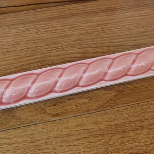 May include: A rectangular, pink ceramic tile with a braided design. The tile has a glossy finish and is set against a wooden surface. The pink color is consistent throughout the braided pattern.