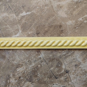 May include: A pale yellow ceramic tile with a rope-like design. The tile is rectangular and features a repeating pattern of curved, wave-like shapes along its length. This decorative tile is suitable for various home decor projects.
