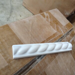 May include: A white, rectangular decorative molding with a rope-like design. The molding is made of a smooth, glossy material and is likely used for interior design or architectural detailing. It is resting on a brown cardboard surface.