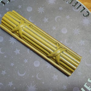 May include: A rectangular, olive-green ceramic object with a fluted, column-like texture featuring two horizontal bands that cross over the vertical ridges. The piece has a glossy finish and is positioned diagonally across a circular gray surface.