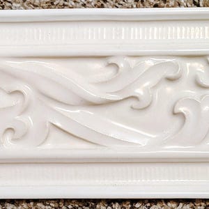May include: A white decorative molding with an ornate, flowing scrollwork design. The molding has a rectangular shape with a detailed relief pattern of stylized leaves and curves. The top and bottom edges have a simple, linear design.