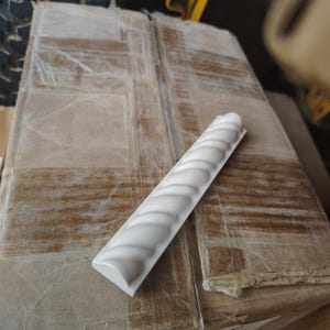 May include: A white, decorative tile with a repeating, curved design. The tile is rectangular and has a glossy finish. It rests on a cardboard box, suggesting it's part of a larger shipment of building materials.