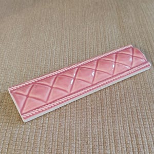 May include: A pink ceramic tile with a diamond pattern and a rope-like border. The tile is rectangular with a curved edge and a glossy finish. The tile is likely for use in a kitchen or bathroom.