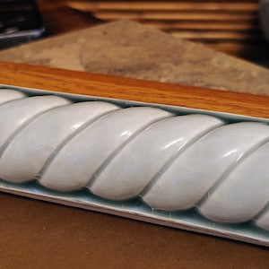 May include: A rectangular ceramic tile featuring a raised, light blue, rope-like twisted design with a glossy, crackled glaze finish. The tile has a smooth, rounded texture and is positioned horizontally, showcasing its repeating spiral pattern.